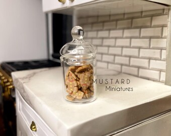 May include: A clear glass jar filled with gingerbread cookies. The jar has a lid and is sitting on a white countertop. The text "MUSTARD Miniatures" is visible in the image.