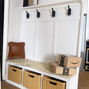 May include: Miniature white entryway bench with three woven baskets, black hooks, and a shelf. Includes a brown pillow, two Amazon boxes, a plant, and yellow clogs. The bench is a dollhouse-sized item.