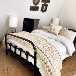 May include: A black metal bed frame with a white duvet and a beige knitted throw blanket. The bed is in a room with a white wall and a wooden floor. The wall has three decorative keyhole-shaped signs that say "Laugh", "Dream", and "Key".