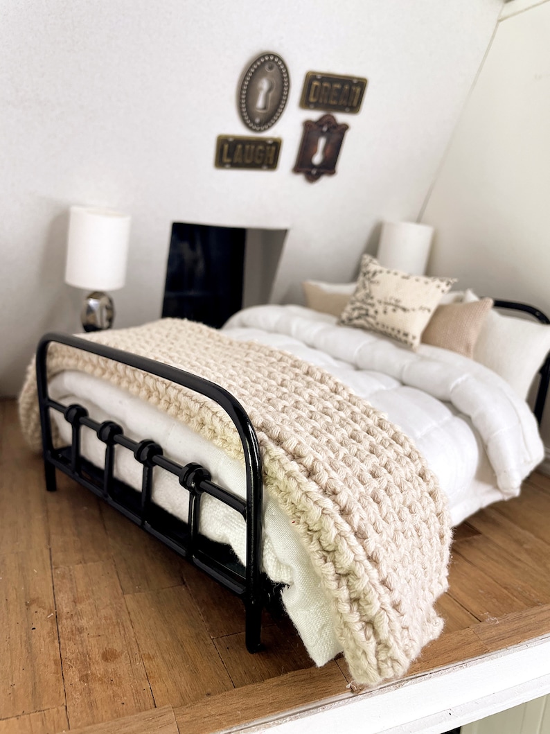 May include: A black metal bed frame with a white duvet and a beige knitted throw blanket. The bed is in a room with a white wall and a wooden floor. The wall has three decorative keyhole-shaped signs that say "Laugh", "Dream", and "Key".