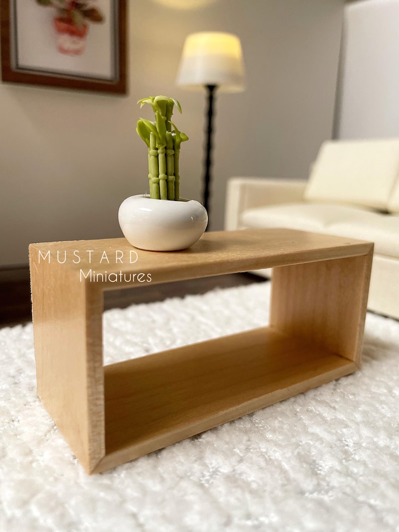 May include: A miniature wooden coffee table with a light brown finish. A small white ceramic pot with a green bamboo plant sits on top. The text "MUSTARD Miniatures" is visible on the table.