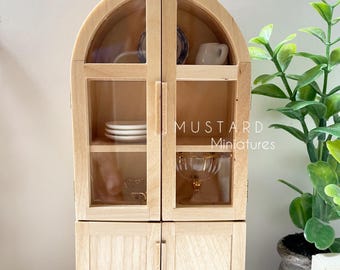 May include: A miniature wooden cabinet with an arched top and glass doors. Inside are small white plates, a teacup, and a decorative glass bowl. The cabinet is light-colored wood and has the text "MUSTARD Miniatures".