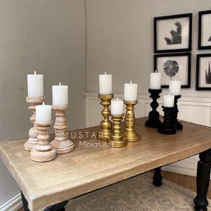May include: Miniature candle holders in various styles, including gold, black, and distressed wood, each holding a white candle. The holders are arranged on a wooden table with black legs, creating a cozy, decorative scene. The text "MUSTARD Miniatures" is visible.