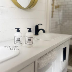 May include: A miniature bathroom scene featuring two white bottles labeled "Hand Lotion" and "Hand Soap" with black pumps. The bottles sit on a white countertop with a black faucet and a gold-framed mirror. The scene is from Mustard Miniatures.