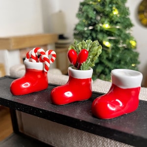 May include: Three red and white ceramic Christmas boot ornaments. The boot on the left is filled with candy canes, the middle boot is filled with red berries and greenery, and the boot on the right is empty.