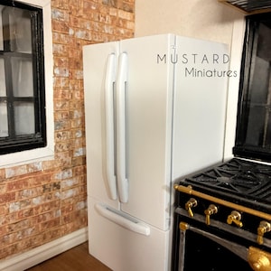Op de afbeelding: Een witte miniatuurkoelkast met twee verticale deuren en een onderste vrieslade, voorzien van lange handgrepen. De tekst MUSTARD Miniatures staat rechtsboven op het apparaat gedrukt, dat naast een zwart fornuis is geplaatst.