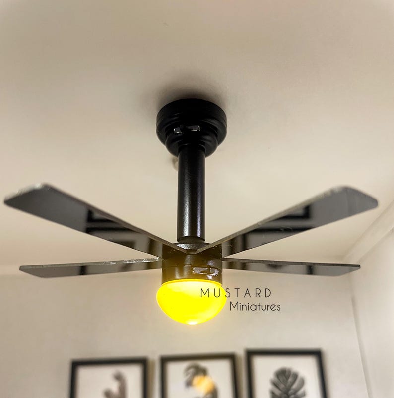 Puede incluir: Un ventilador de techo negro en miniatura con cinco aspas y una l&aacute;mpara central. La luz emite un brillo amarillo c&aacute;lido. El texto "MUSTARD Miniatures" est&aacute; impreso en la l&aacute;mpara.