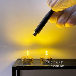 Puede incluir: Dos pequeñas luces LED doradas con un brillo amarillo cálido. Las luces están sobre una superficie negra, con el texto "MUSTARD Miniatures" visible. Un cable negro se sostiene sobre las luces.