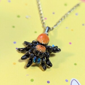 Pode incluir: Um colar de corrente de prata com um pingente de aranha preto e laranja. A aranha tem olhos azuis e é detalhada com esmalte preto e laranja.