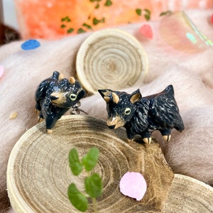 May include: A pair of black ceramic goat earrings with gold accents. The earrings are displayed on a wooden surface with a green leaf and a pink flower.