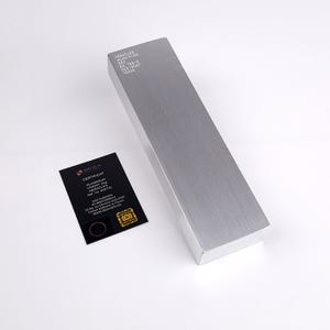May include: A silver aluminum ingot, rectangular in shape, with the text "HERACLES ALUMINIUM". A black certificate card with gold detailing lies beside the ingot. The ingot's dimensions are not visible.