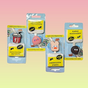 May include: Three colorful packaging displays for phone cable accessories. Each display features a different cartoon character: a soda can, a pink character with a bow, and a black character with a white face. The text on the packaging reads "Data Line Protection", "Cable Accessory", and "Cable Accessory".