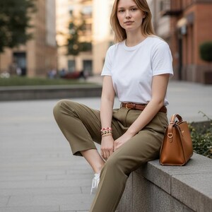 May include: A woman wearing a white t-shirt, olive green pants, and white sneakers. She is also wearing a brown belt and a bracelet. A brown leather handbag sits beside her. The background features a city street with buildings.