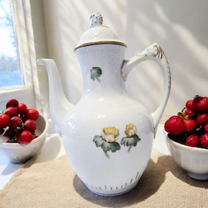 Puede incluir: Cafetera de porcelana blanca con tapa, boquilla y asa. La cafetera está decorada con adornos dorados y diseños florales. Dos pequeños cuencos de bayas rojas están a cada lado.