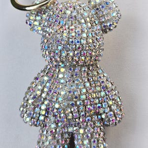 以下が含まれることがあります： キラキラ光るラインストーンのテディベアのキーホルダー。金色の留め金が付いています。クマは小さくカラフルなラインストーンで覆われており、きらびやかな効果を生み出しています。キュートで華やかなアクセサリーです。
