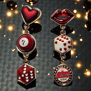 May include: Dangling earrings with a gold finish. One earring has a red heart, a red 7-ball, and a red dice with white hearts. The other has red lips, a white and red heart dice, and a Cherry Soda emblem.