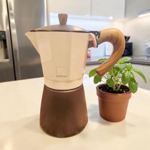 Puede incluir: Una cafetera italiana con la parte superior color crema, base marrón oscuro y un asa y pomo de tapa con patrón de vetas de madera. Un pequeño detalle con la bandera italiana está unido al asa. Al lado hay una maceta de terracota con una planta de albahaca.