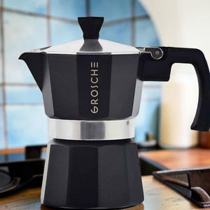 May include: A black moka pot coffee maker with a faceted octagonal body and a silver-colored middle band. The word GROSCHE is printed vertically in white on the upper chamber. It features a black handle, a black lid knob, and a silver-colored lid.
