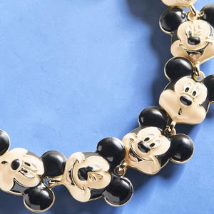 May include: A gold-toned necklace featuring multiple Mickey Mouse face charms with black and cream enamel detailing. The necklace has a classic design with a playful and recognizable character.