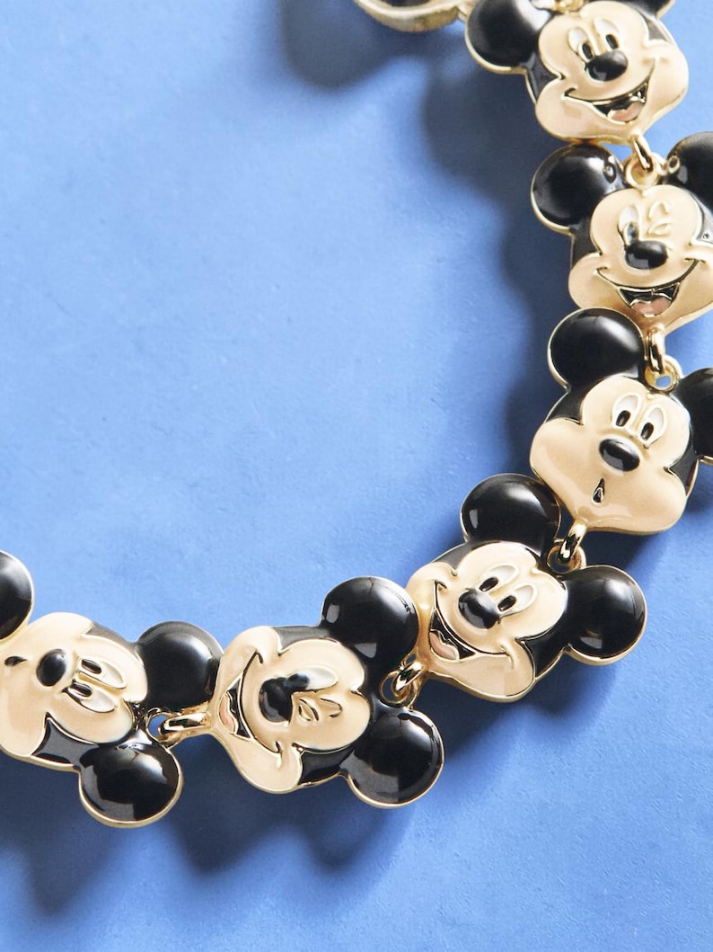 May include: A gold-toned necklace featuring multiple Mickey Mouse face charms with black and cream enamel detailing. The necklace has a classic design with a playful and recognizable character.