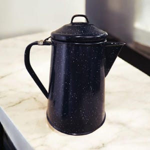 May include: A black enamel coffee pot with a lid and handle. The pot has a speckled white pattern and a spout for pouring. The pot is sitting on a white marble surface. The coffee pot is a kitchen item.