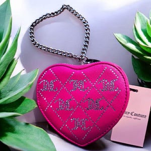 May include: A vibrant pink heart-shaped purse with a silver chain strap. The purse is decorated with a diamond pattern and silver rhinestone "JC" lettering. A tag with "Juicy Couture" is attached.