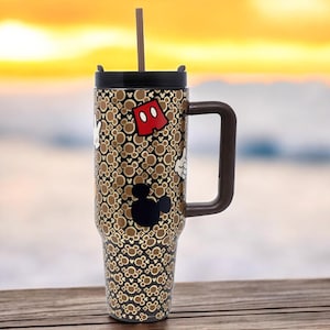 K&ouml;nnte beinhalten: Ein Edelstahl-Reisebecher mit braunem Griff und Deckel. Der Becher hat ein Muster aus Mickey Mouse-Symbolen und ein braunes und wei&szlig;es Design. Ein brauner Strohhalm ist in den Deckel gesteckt. Der Becher steht auf einer Holzoberfl&auml;che.