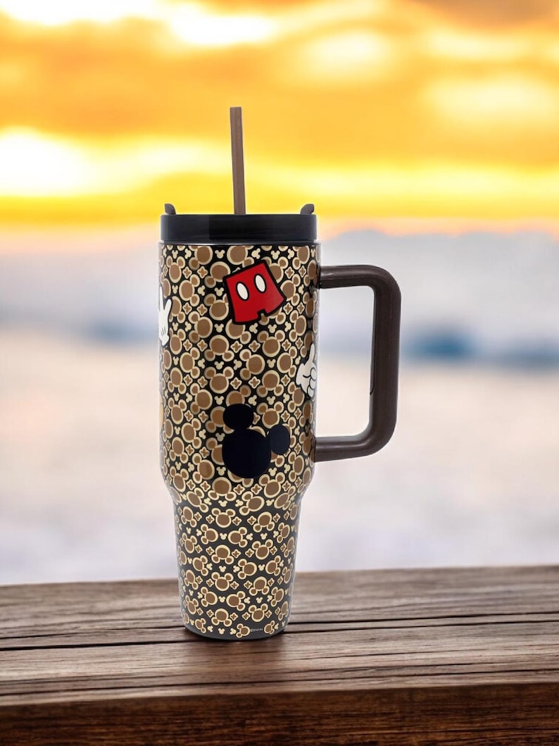 K&ouml;nnte beinhalten: Ein Edelstahl-Reisebecher mit braunem Griff und Deckel. Der Becher hat ein Muster aus Mickey Mouse-Symbolen und ein braunes und wei&szlig;es Design. Ein brauner Strohhalm ist in den Deckel gesteckt. Der Becher steht auf einer Holzoberfl&auml;che.