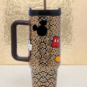 K&ouml;nnte beinhalten: Ein Edelstahl-Reisebecher mit einem braunen und schwarzen Mickey Mouse-Muster. Der Becher hat einen schwarzen Deckel, einen braunen Griff und einen Strohhalm. Mickeys Silhouette, Handschuh, Shorts und ein gelber Knopf sind abgebildet.