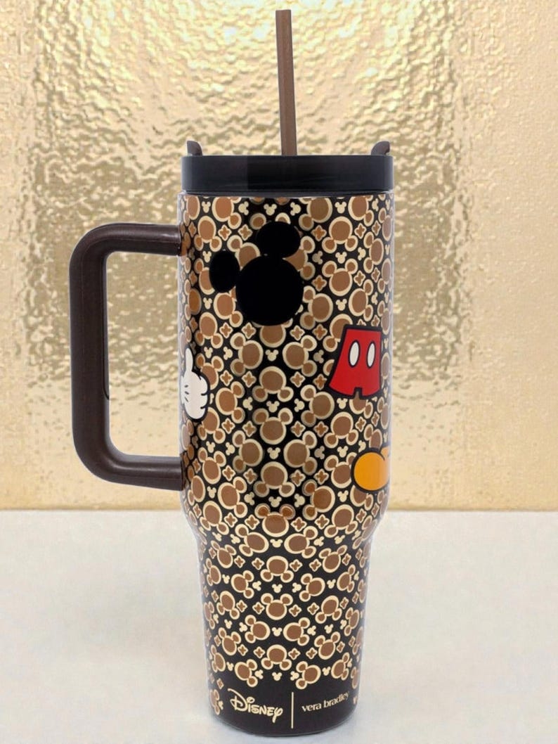 K&ouml;nnte beinhalten: Ein Edelstahl-Reisebecher mit einem braunen und schwarzen Mickey Mouse-Muster. Der Becher hat einen schwarzen Deckel, einen braunen Griff und einen Strohhalm. Mickeys Silhouette, Handschuh, Shorts und ein gelber Knopf sind abgebildet.