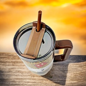 K&ouml;nnte beinhalten: Ein brauner und durchsichtiger Reisebecher mit Henkel und Deckel. Der Becher hat einen braunen Strohhalm und ein Design mit einer rot-wei&szlig;en Figur. Der Becher steht auf einer Holzoberfl&auml;che.