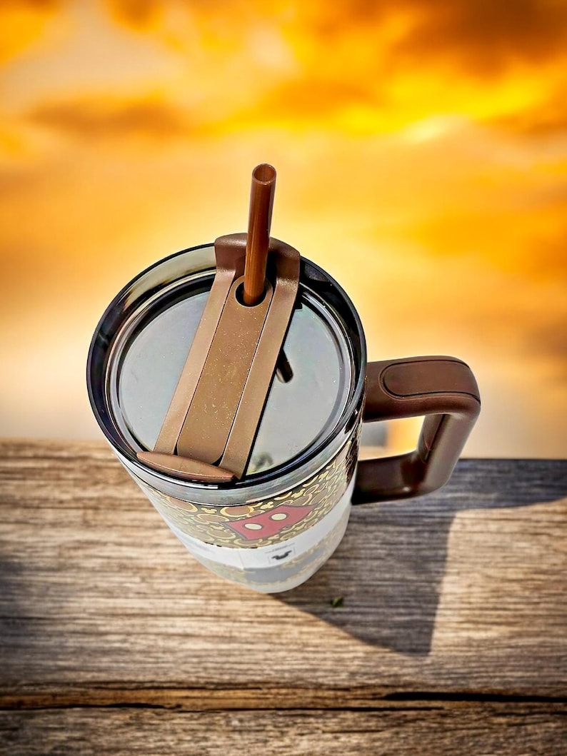 K&ouml;nnte beinhalten: Ein brauner und durchsichtiger Reisebecher mit Henkel und Deckel. Der Becher hat einen braunen Strohhalm und ein Design mit einer rot-wei&szlig;en Figur. Der Becher steht auf einer Holzoberfl&auml;che.