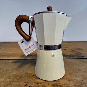 May include: A cream-colored moka pot with a wood-grain patterned handle and lid knob. The coffee maker features a metallic silver band around the center and a small gold-toned valve. A white tag attached to the handle reads gnali&zani.
