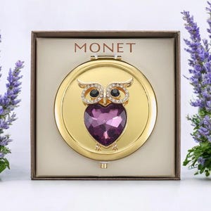 May include: A gold-toned compact mirror featuring an owl design. The owl has crystal accents and black eyes, with a heart-shaped purple gem. The compact is in a brown box with the brand name "MONET" printed on it.