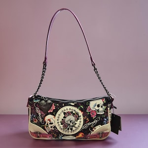 Könnte beinhalten: Schultertasche mit schwarzem Blumenmuster, lila Riemen und Kette. Die Tasche hat Totenkopf- und Rosenmotive, einen cremefarbenen Boden und den Text "Ed Hardy". Ein schwarzes Etikett ist angebracht.