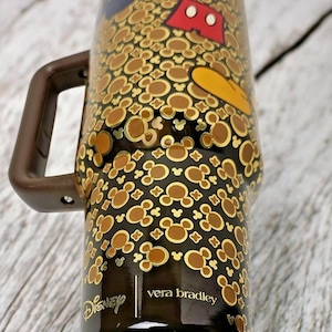 K&ouml;nnte beinhalten: Ein schwarz-brauner Reisebecher mit Henkel, der ein Muster aus Mickey Mouse-Silhouetten und Charakterdetails aufweist. Der Becher hat die Logos von Disney und Vera Bradley.