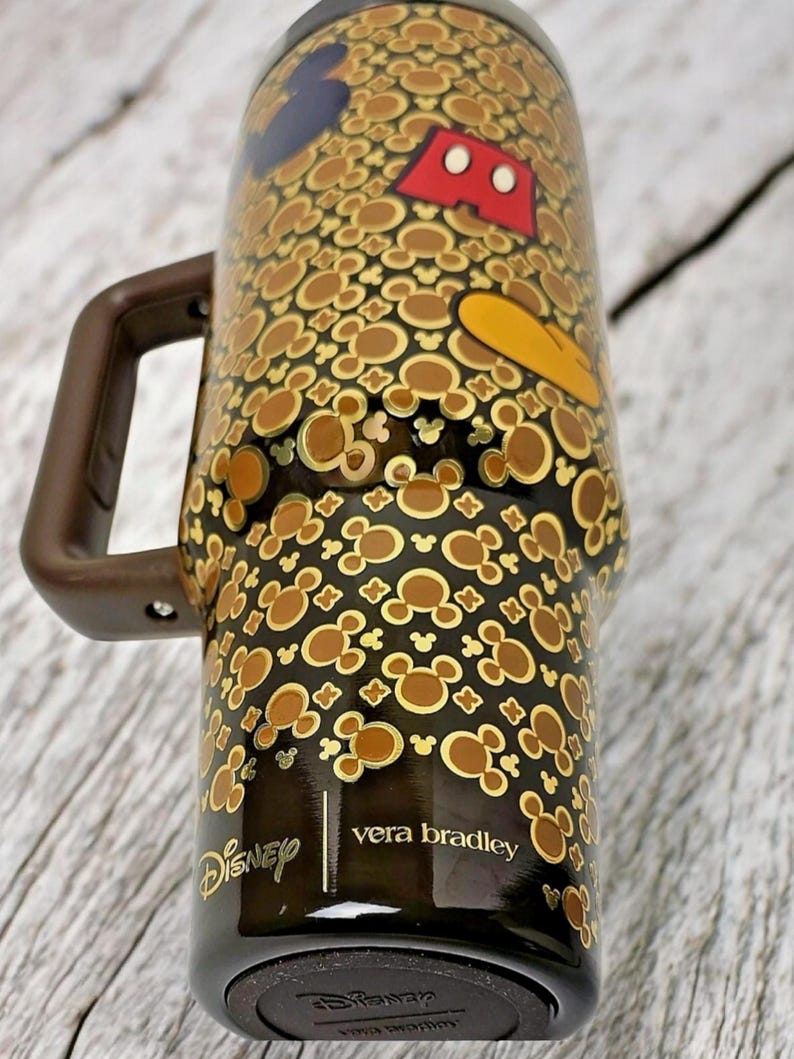K&ouml;nnte beinhalten: Ein schwarz-brauner Reisebecher mit Henkel, der ein Muster aus Mickey Mouse-Silhouetten und Charakterdetails aufweist. Der Becher hat die Logos von Disney und Vera Bradley.