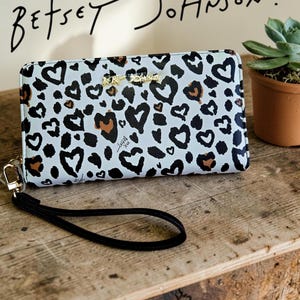 Op de afbeelding: Een witte portemonnee met een zwart en bruin hartvormig luipaardprintpatroon. De portemonnee heeft een zwart polsbandje en goudkleurige hardware. De merknaam "Betsey Johnson" staat in zwart schrift boven de portemonnee.