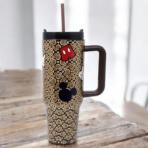 K&ouml;nnte beinhalten: Ein hoher Reisebecher mit braunem Griff und Deckel, mit einem sich wiederholenden Muster von Mickey Mouse-Symbolen in Braun und Gold. Der Becher hat eine schwarze Mickey Mouse-Silhouette und eine rote Shorts-Grafik. Ein brauner Strohhalm ragt aus dem Deckel.