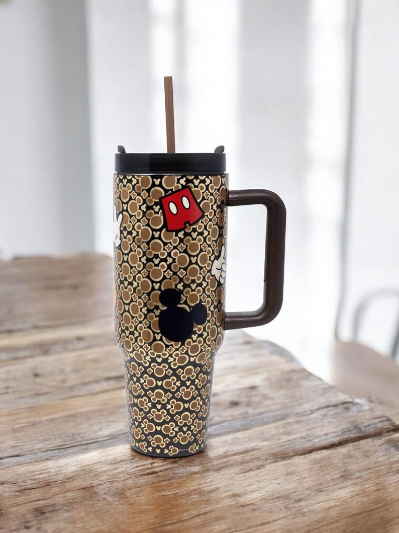 K&ouml;nnte beinhalten: Ein hoher Reisebecher mit braunem Griff und Deckel, mit einem sich wiederholenden Muster von Mickey Mouse-Symbolen in Braun und Gold. Der Becher hat eine schwarze Mickey Mouse-Silhouette und eine rote Shorts-Grafik. Ein brauner Strohhalm ragt aus dem Deckel.