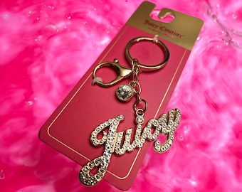 May include: A gold-colored keychain with the word "Juicy" spelled out in sparkling rhinestones. The keychain is attached to a red card with the "Juicy Couture" logo. The background is a vibrant pink.