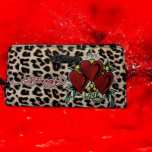May include: Leopard print wallet with the Ed Hardy logo and a tattoo-style design of three red hearts with the word "LOVE". The wallet has a black zipper and is set against a red background.