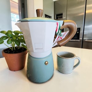 May include: A white and teal stovetop espresso maker with a wooden handle and lid knob. A small ceramic mug and a potted plant are next to the coffee maker. The text "grab & cool" is visible on the coffee maker.