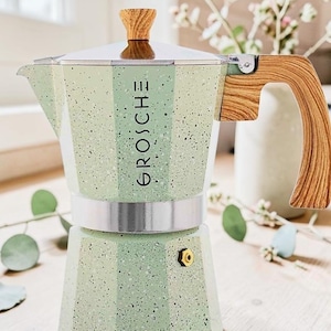 May include: A light green stovetop espresso maker with a speckled finish and the word GROSCHE printed vertically on the side. It features a metallic silver middle band, a wood-grain patterned handle, and a matching wood-grain knob on the lid.