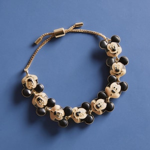 May include: Gold-toned bracelet featuring Mickey Mouse charms with black ears and faces. The adjustable bracelet has a sliding clasp and is set against a blue background. The charms are evenly spaced along the chain.