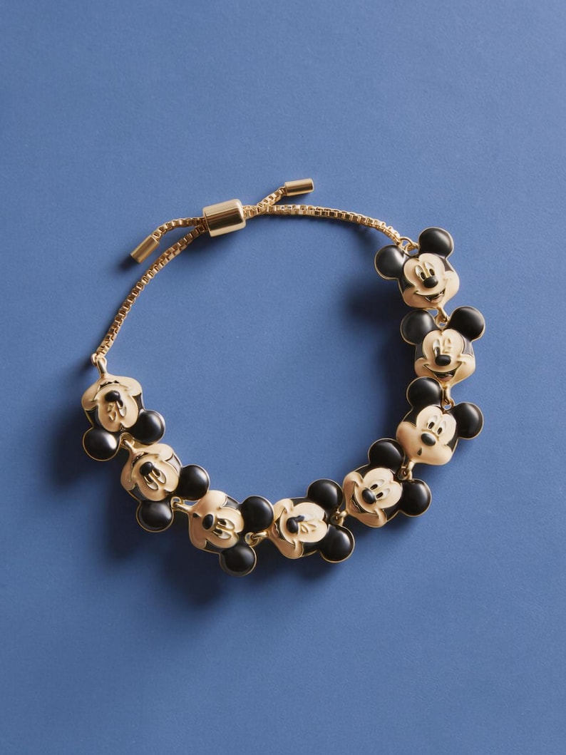 May include: Gold-toned bracelet featuring Mickey Mouse charms with black ears and faces. The adjustable bracelet has a sliding clasp and is set against a blue background. The charms are evenly spaced along the chain.