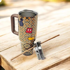 K&ouml;nnte beinhalten: Ein hoher Reisebecher mit braunem Griff und Deckel, verziert mit Mickey Mouse Silhouetten und Icons in Braun, Beige und Schwarz. Der Becher enth&auml;lt einen Strohhalm und steht auf einer Holzoberfl&auml;che.