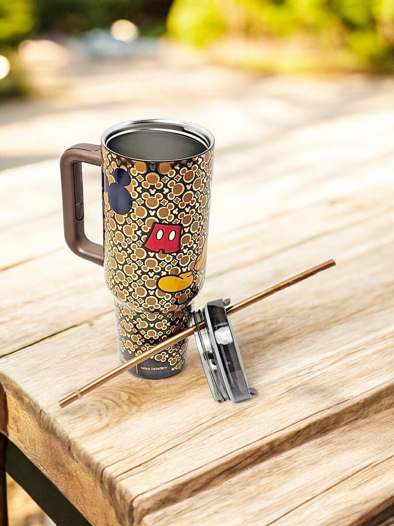 K&ouml;nnte beinhalten: Ein hoher Reisebecher mit braunem Griff und Deckel, verziert mit Mickey Mouse Silhouetten und Icons in Braun, Beige und Schwarz. Der Becher enth&auml;lt einen Strohhalm und steht auf einer Holzoberfl&auml;che.