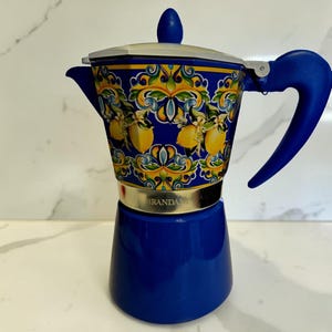May include: A blue stovetop espresso maker with a decorative floral and lemon design. The handle and lid are blue, and the base is a solid blue color. The espresso maker has a silver band around the middle and the brand name is visible.