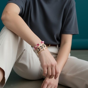May include: A person wearing a dark gray t-shirt and light beige pants. The person is wearing three bracelets: one with pink beads, one with black flower-shaped beads, and a gold bracelet with a pearl.